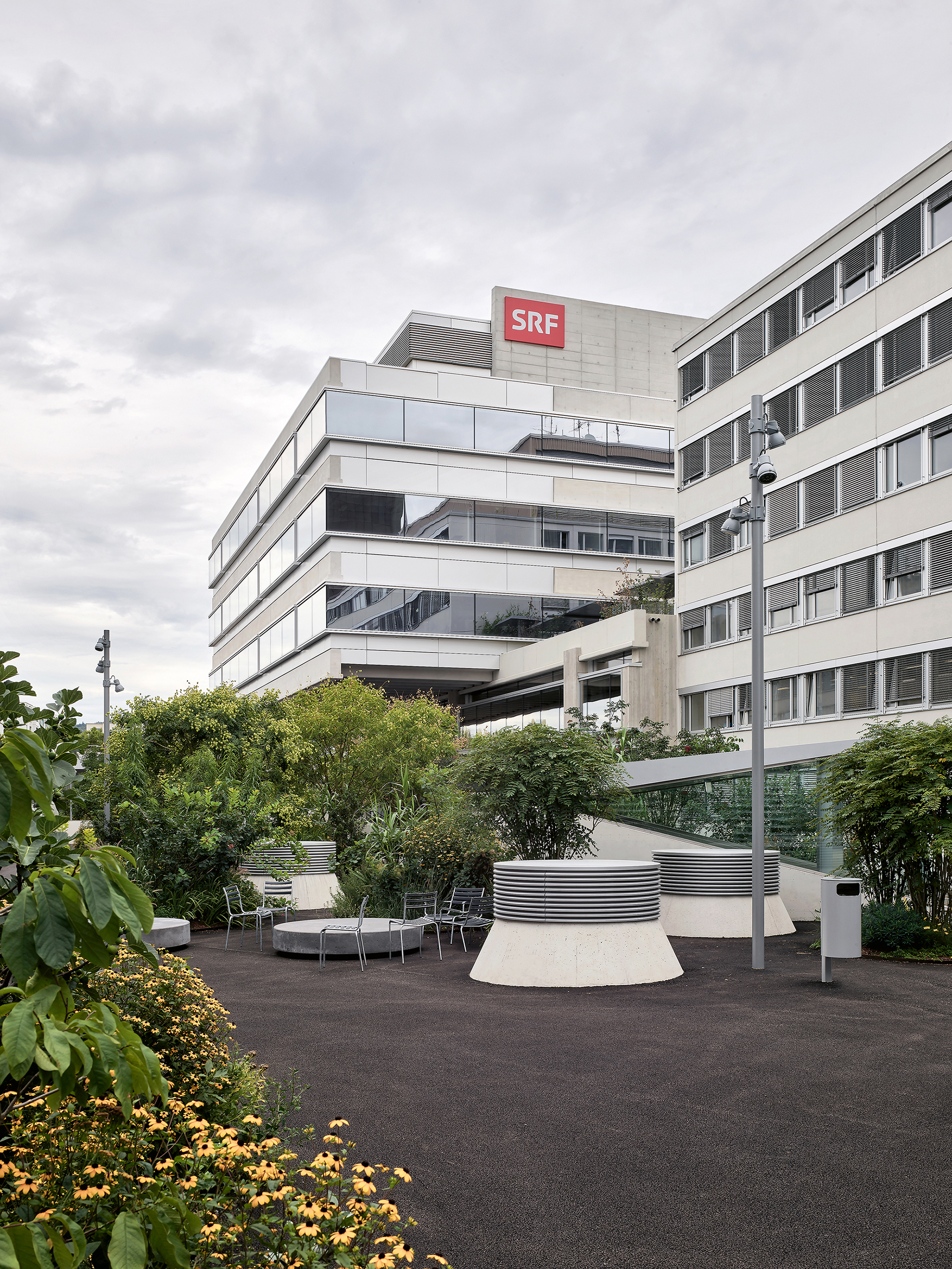 Der Aussenraum ist als öffentlicher Park gestaltet: Grüne Inseln unterschiedlicher Grösse bieten im «Mediengarten» verschiedene Grade  an Intimität.  Die offenen Rampenaugen schaffen eine Sichtverbindung zum darunter liegenden Parkhaus, Lüftungs- und Entrauchungs- auslässe sind in den Garten integriert.