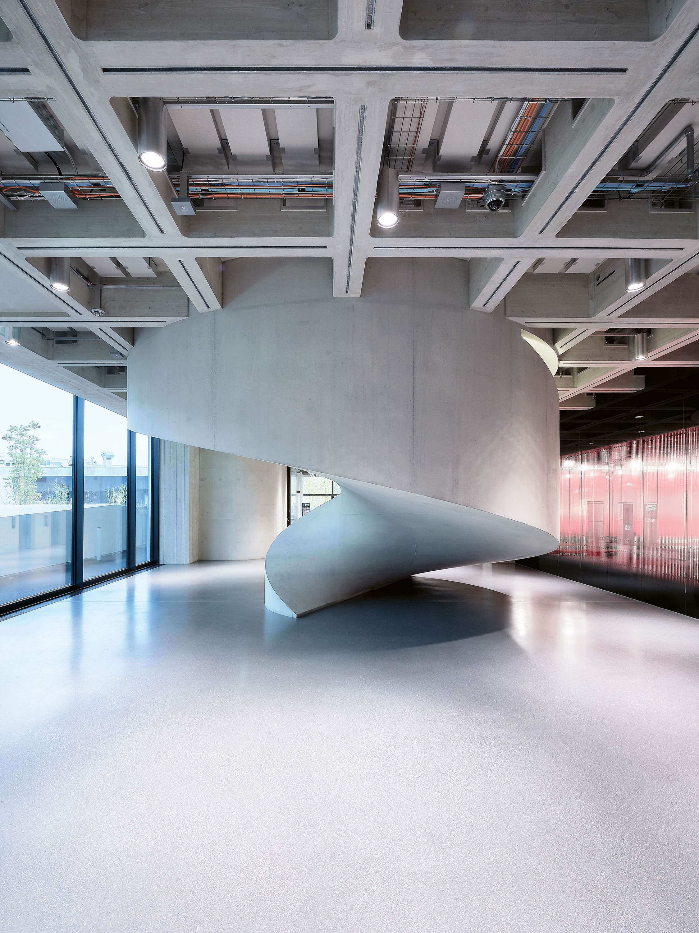 Vollendete Eleganz, klare Funktion, rohes Material: Die Wendeltreppe bringt das architektonische Konzept des Neubaus auf den Punkt.  In der Eingangshalle im EG kommt sie spektakulär auf den Boden, empfängt die Eintretenden wie ein plastisches Kunstwerk und verleiht dem nüchternen Raum einen repräsentativen Charakter. Weil sich das Treppenauge nach oben kontinuierlich ausweitet, wähnt man sich beim Aufstieg im gewundenen Haus einer Meeresschnecke. Links im Bild ist die Fassade zum Mediengarten zu sehen, rech