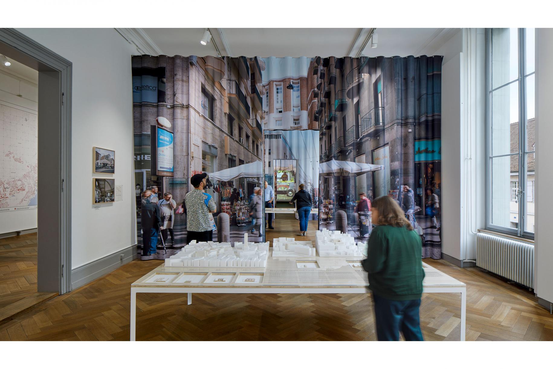 Coup d’œil sur l’exposition: Des étoffes imprimées de motifs photographiques de Naples flottant silencieusement divisent les pièces. Sur les murs, les photos de Cyrille Weiner, sur des tables, des informations détaillées sur les bâtiments. Des «portes» découpées dans les toiles confèrent une vue sur la salle suivante.