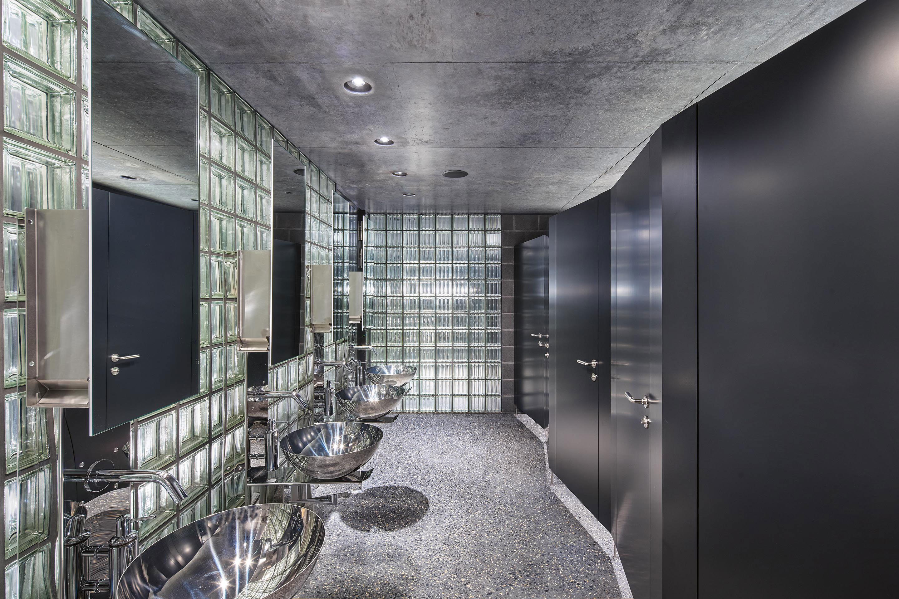 Die Gestaltung der Sanitärräume im Erdgeschoss der Kuben folgt dem industriellen Materialkanon. Eigens angefertigte glänzende Waschplätze werfen Lichtreflexe in den Raum. Als Echo der gefalteten Betonfassade sind auch die innenliegenden Flächen abschnitts­weise geknickt.