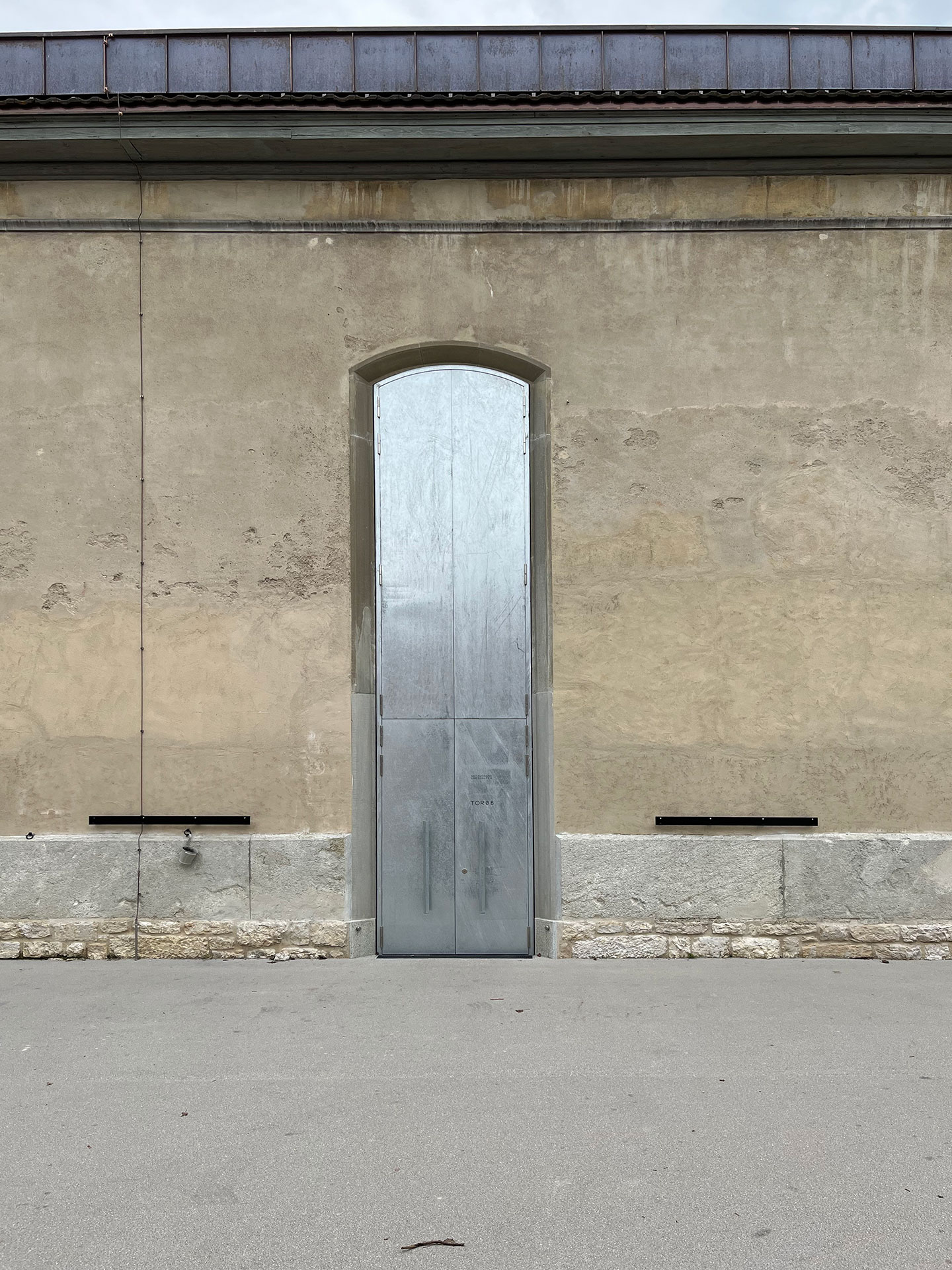 Die mehrschichtige Konstruktion der Fensterläden sichert die Stille im Innenraum. In geöffnetem Zustand gleichen sie überdimensionalen Scheuklappen. Eine mobile Signaletik zeigt den jeweils tagesaktuellen Zugang in die Reithalle an. Dafür werden entsprechend beschriftete Metallbleche in die Bügel, die in der Wand verankert sind, eingehängt.
