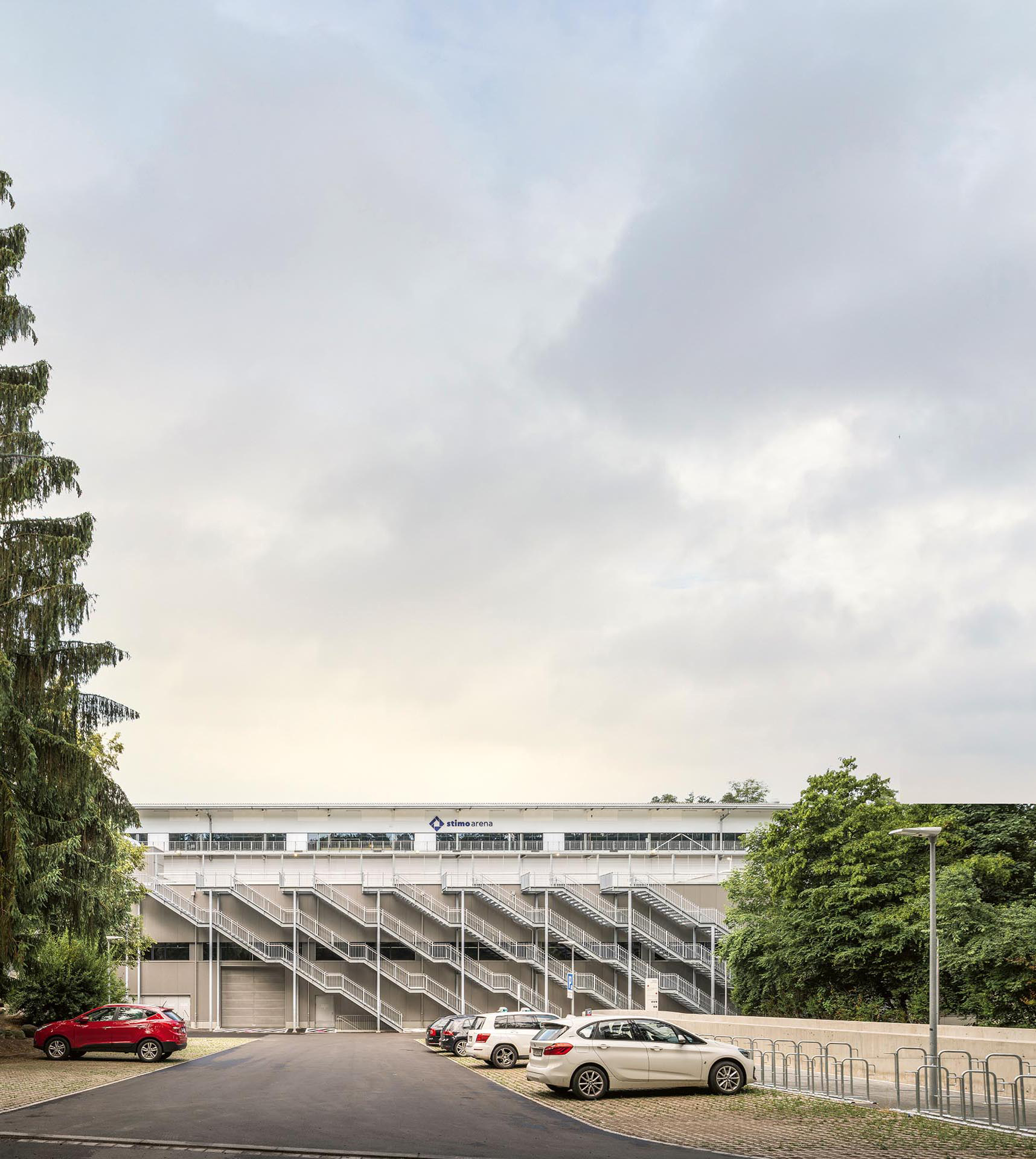Blick vom Schluefweg auf die neue, der Arena vorgelagerten Halle:  Die Dachterrasse ist öffentlich, die Fluchttreppen nutzten die Entwerfenden als üppiges Gestaltungselement für die ansonsten nüchterne Fassade.