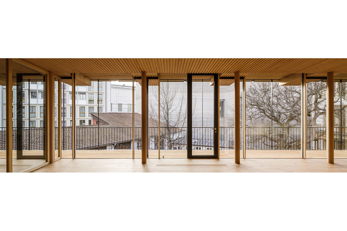 Der Holzbau mit geschlossenen Stirnseiten öffnet sich vom Rand des Grundstücks aus mit einer Glasfassade und  Laubengängen zum Innenhof hin. Gegenüber sieht man den Kunstmuseum-Neubau von Christ  &amp; Gantenbein.