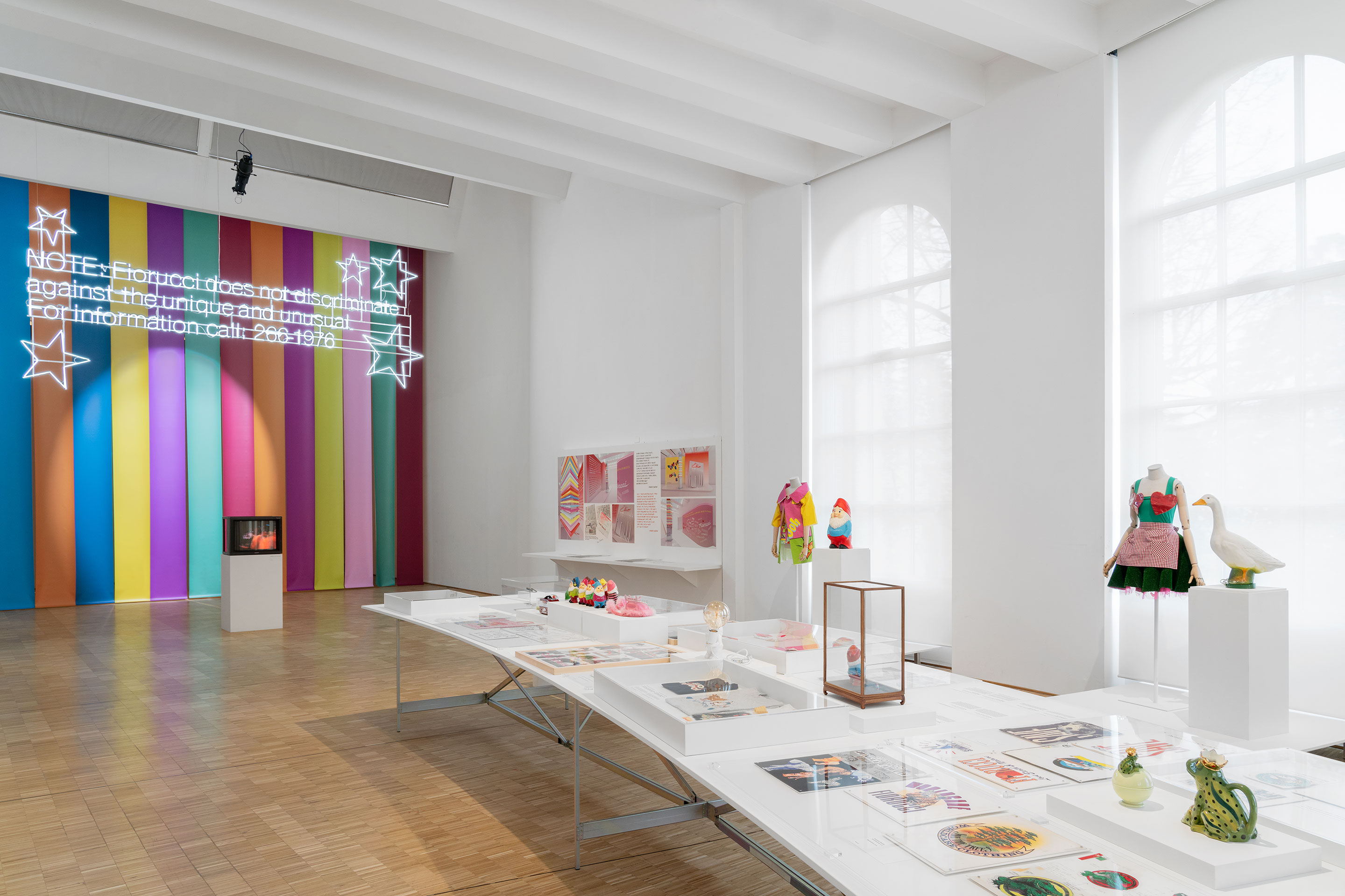 Die Exponate der Schau liegen ausgebreitet auf Tischen oder sind in Bildern dokumentiert, dazu ist die Stimme von Elio Fiorucci zu hören.