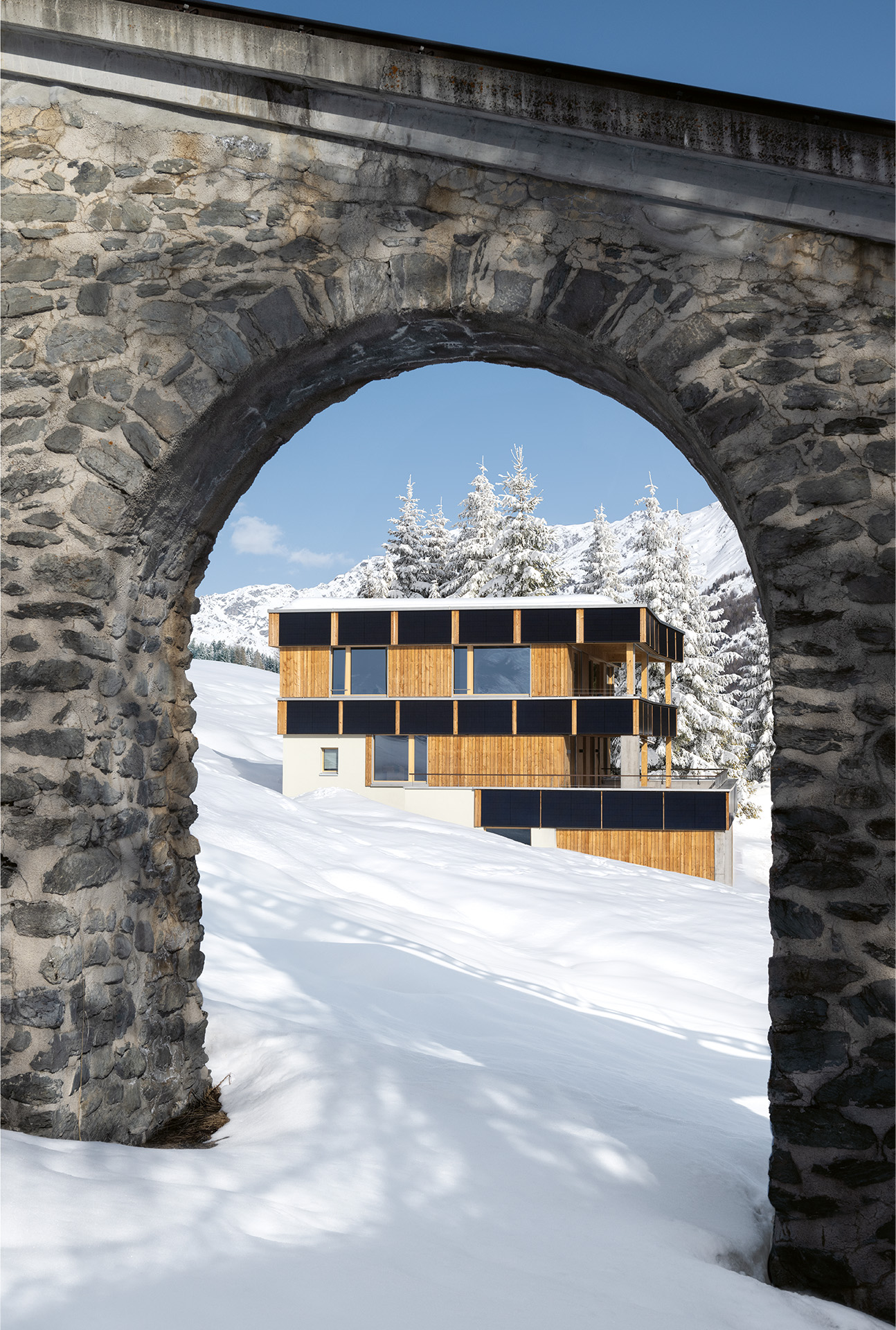 Die Hanglage erforderte einen soliden Sockel aus Beton.  Die Ausrichtung der Wohnungen formte gleichwertige Fassaden. Die schwarzen PV-Bänder unterstreichen die Gestaltung.