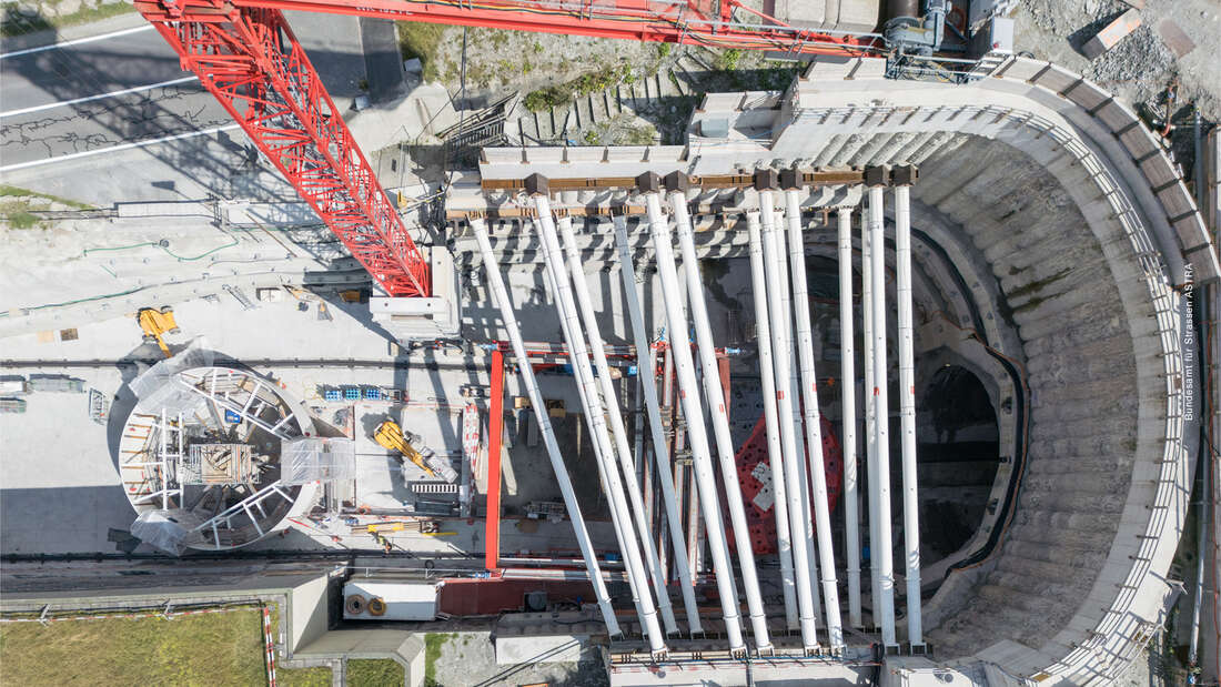Vue aérienne d’un chantier de percement de tunnel: grue rouge, tubes de forage blancs alignés en éventail au-dessus d’une excavation circulaire, structure en béton armé et roue de chantier à droite.