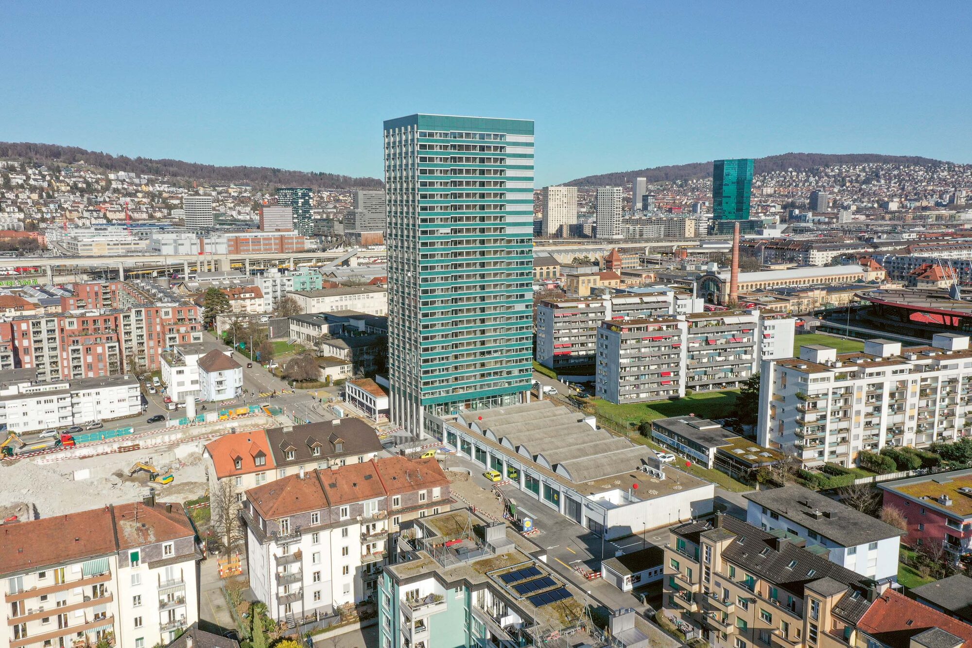 Vue aérienne de l'Alto Tower à Zurich Altstetten, tour de 26 étages entre deux hangars conservés, quartier vu depuis les airs.