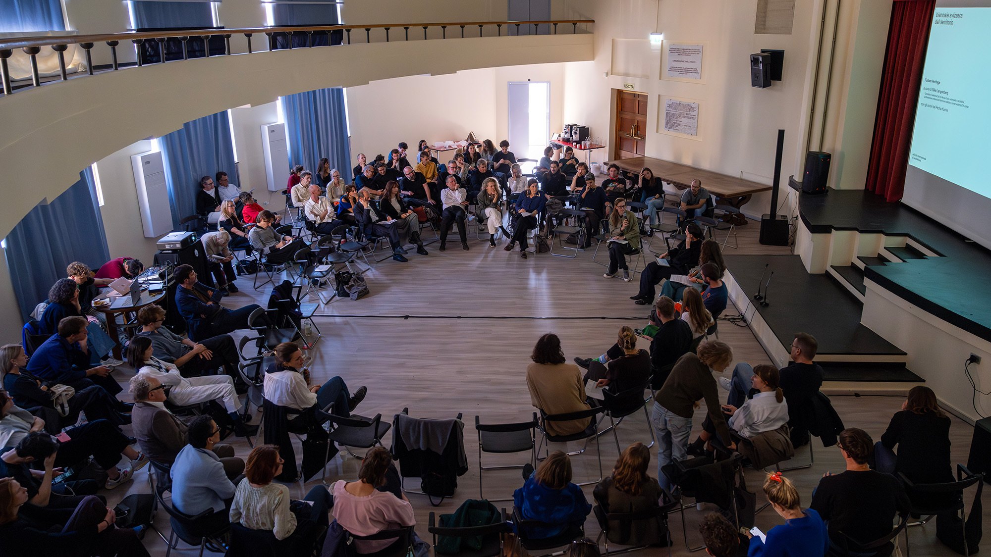 Public assis en cercle dans une salle lors d’un symposium de la biennale suisse du territoire, écran projetant « biennale svizzera del territorio — Future Heritage »