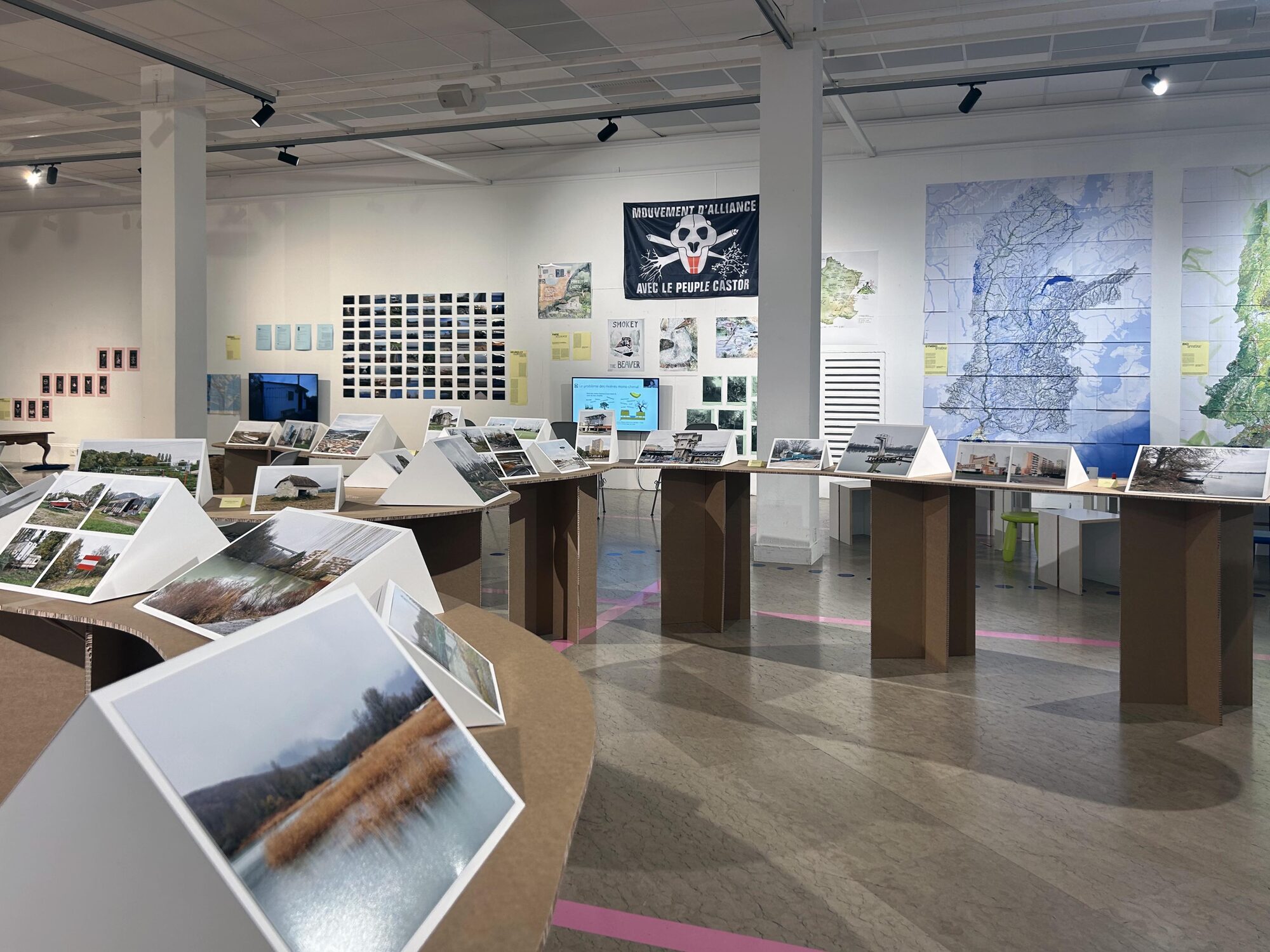 Salle d’exposition du F’AR, tables sinueuses en carton avec pupitres blancs et photographies de paysages.