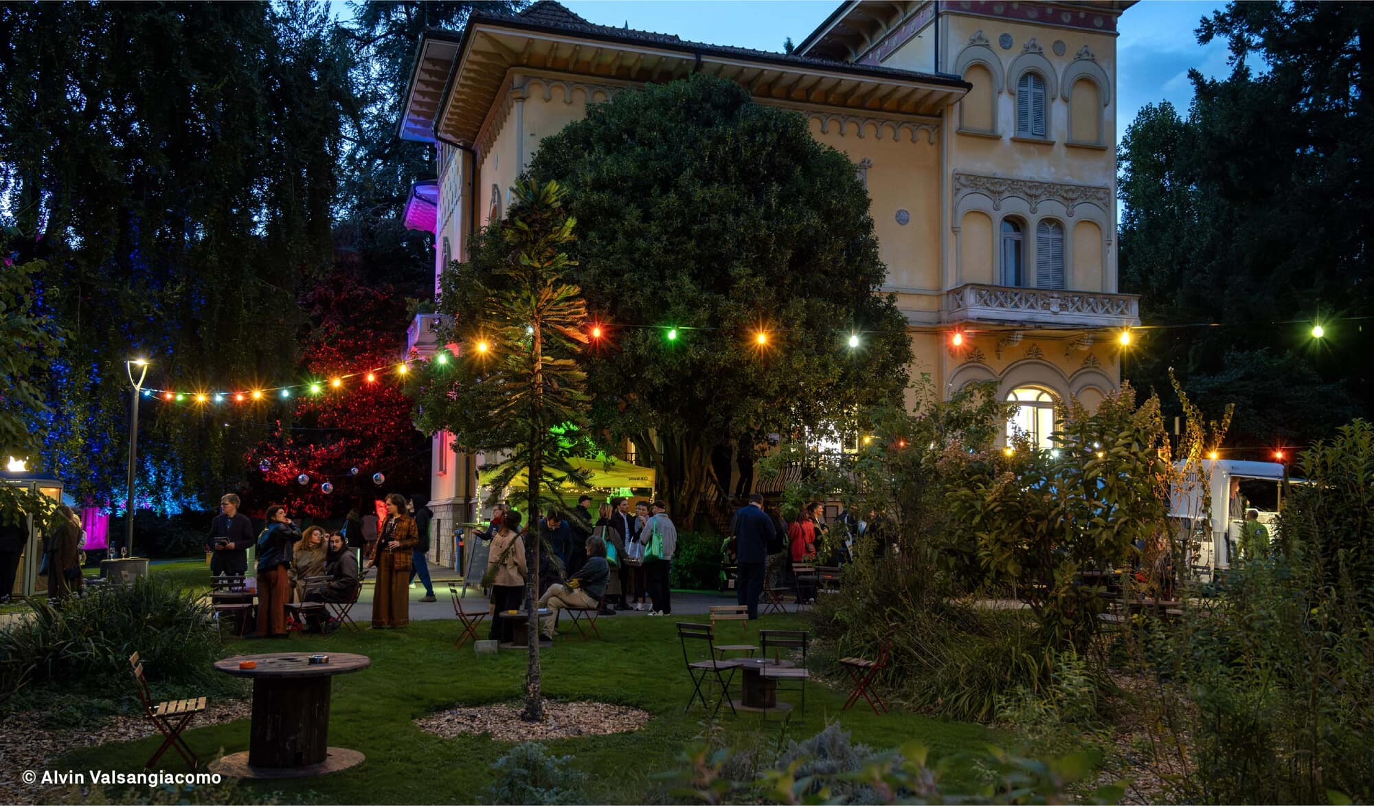 Soirée en plein air dans le parc de Villa Saroli, guirlandes lumineuses entre les arbres, tables et public