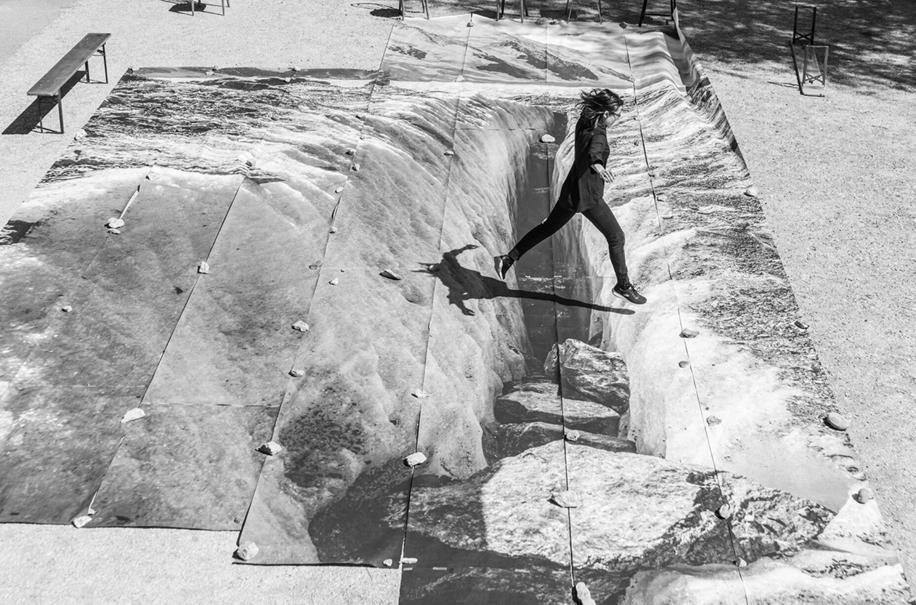 Photographie noir et blanc d’une personne sautant au-dessus d’une installation figurant une crevasse glaciaire au sol, dans un jardin.