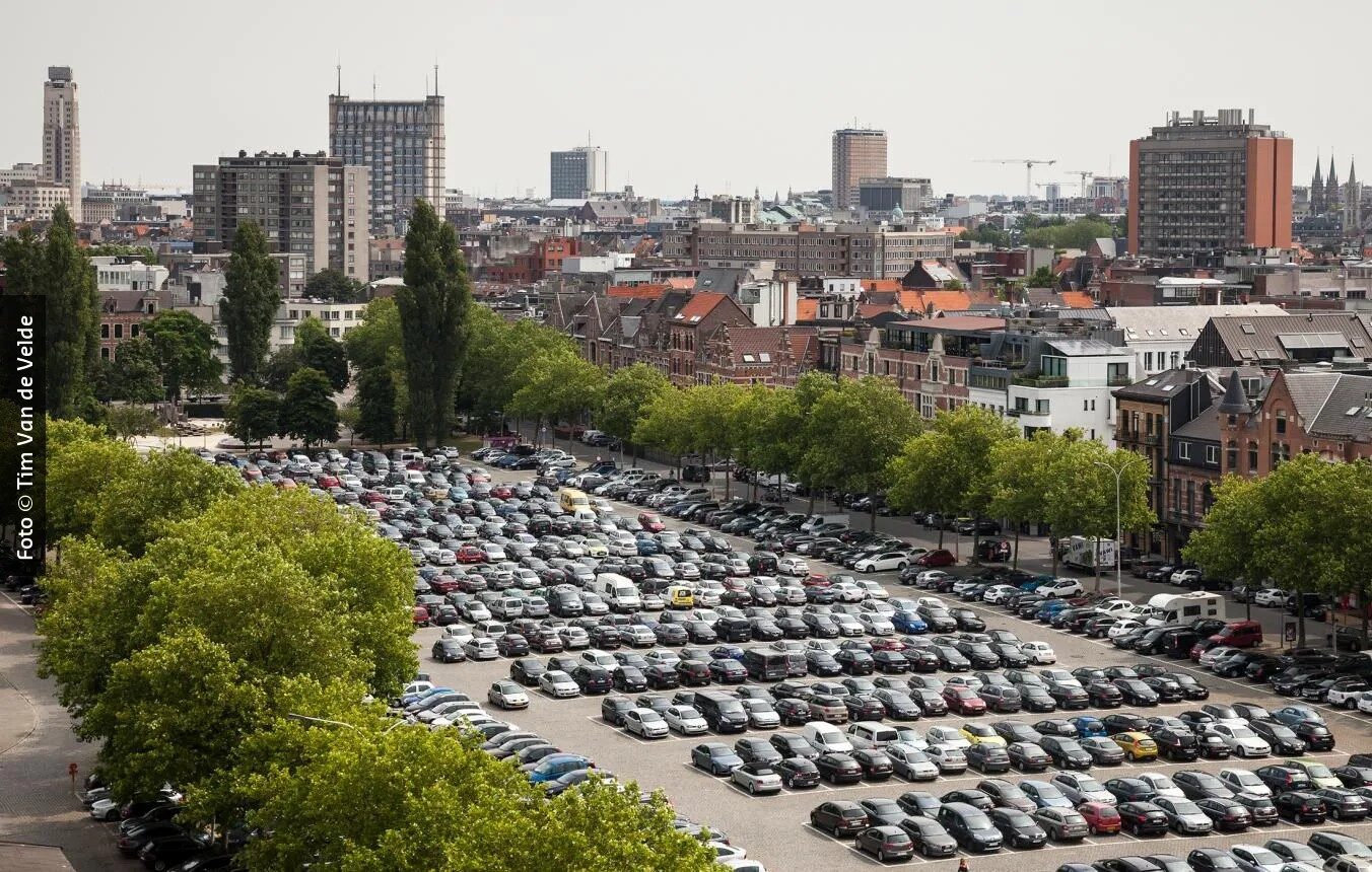 Vue aérienne d’un grand parking rempli de voitures, bordé d’arbres et de bâtiments, à Anvers.