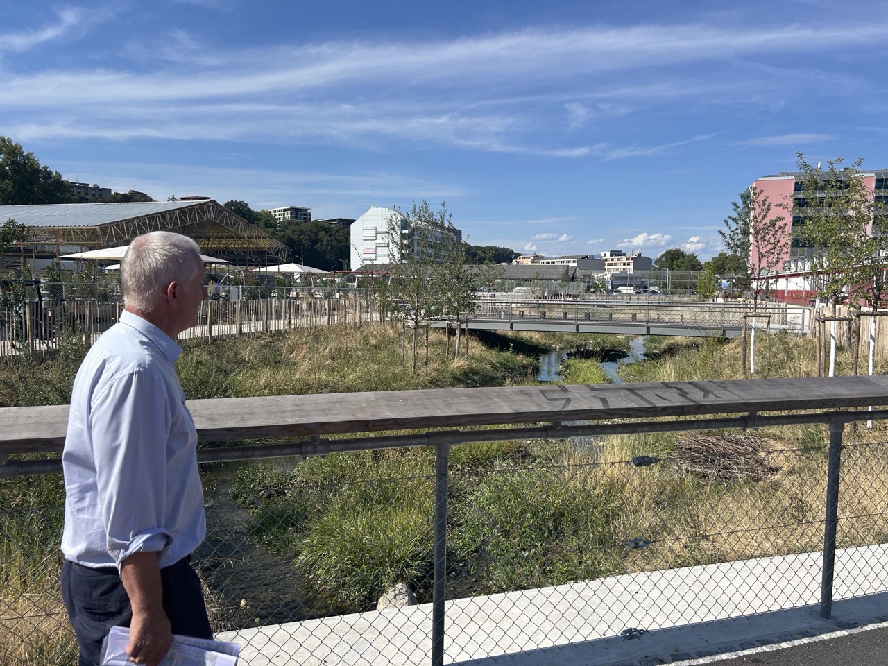 Marco Rampini devant la Drize à ciel ouvert, bordée de hautes herbes, avec une passerelle au fond.