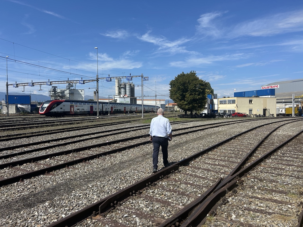 Marco Rampini marche sur des voies ferrées, un train et des halles industrielles en arrière-plan.