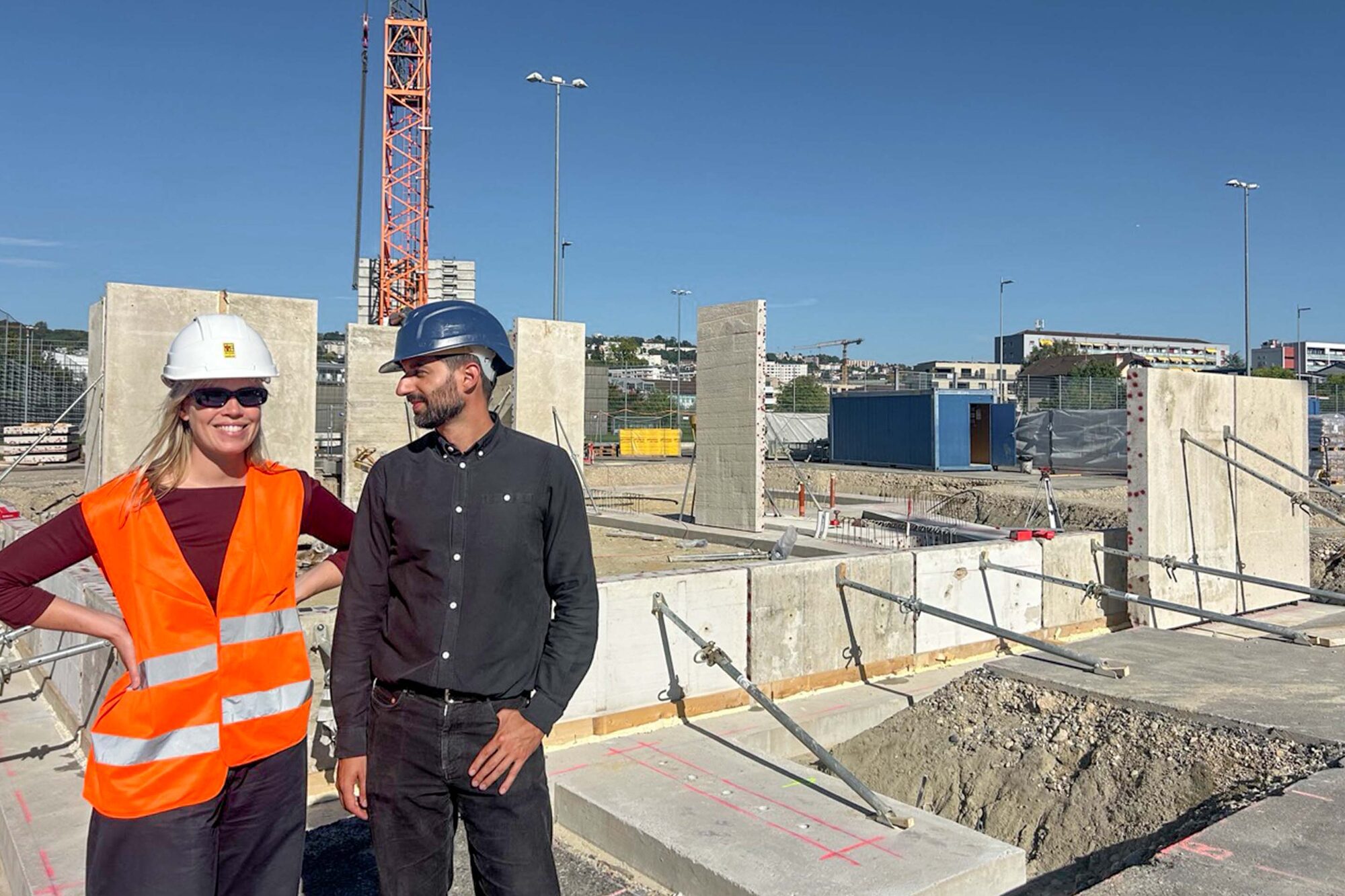 Une femme en gilet orange et casque blanc et un homme en casque bleu posent devant des murs de béton.