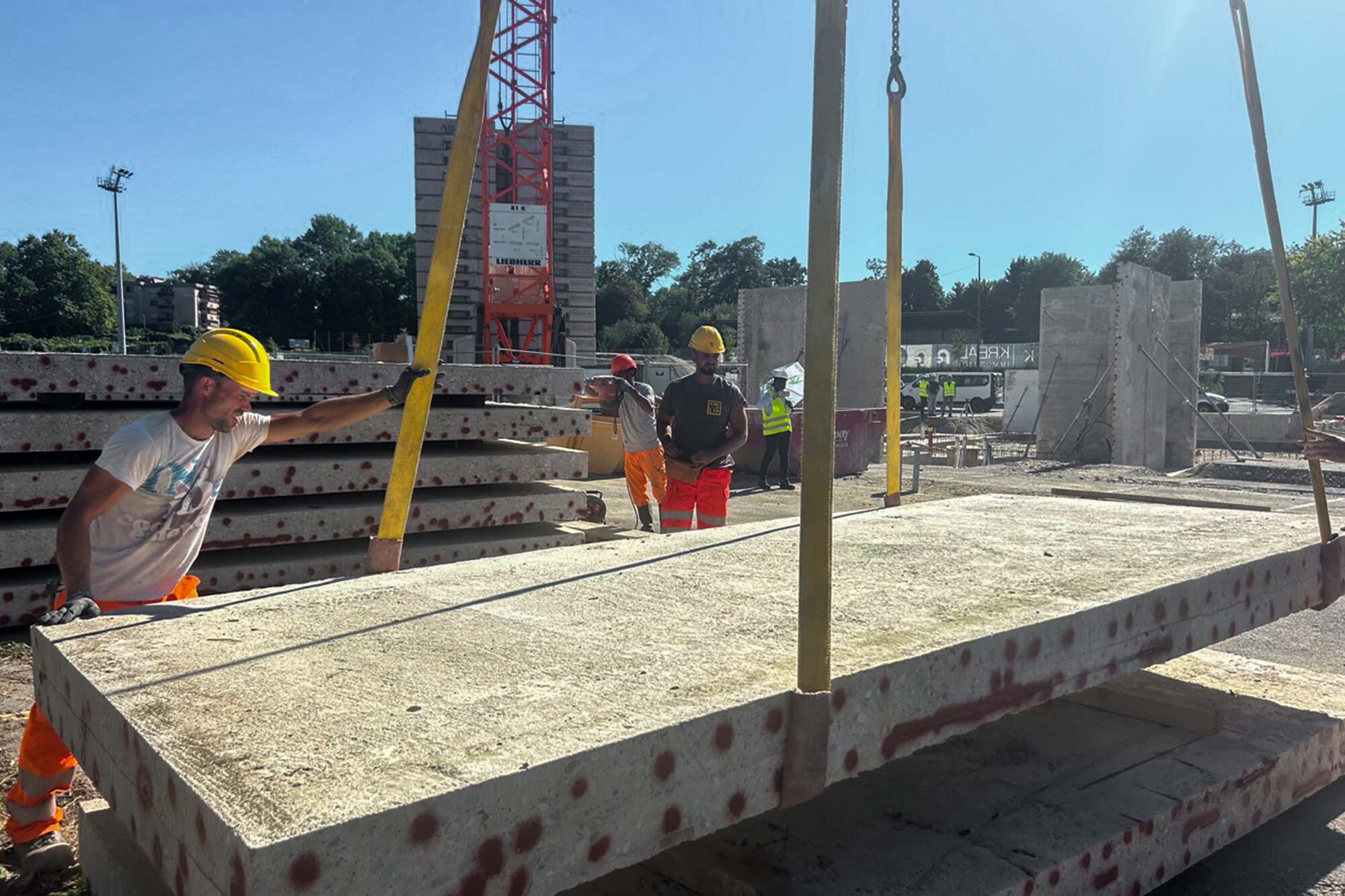 Ouvrier guidant une grande dalle de béton suspendue à une grue, avec d’autres dalles empilées derrière lui.
