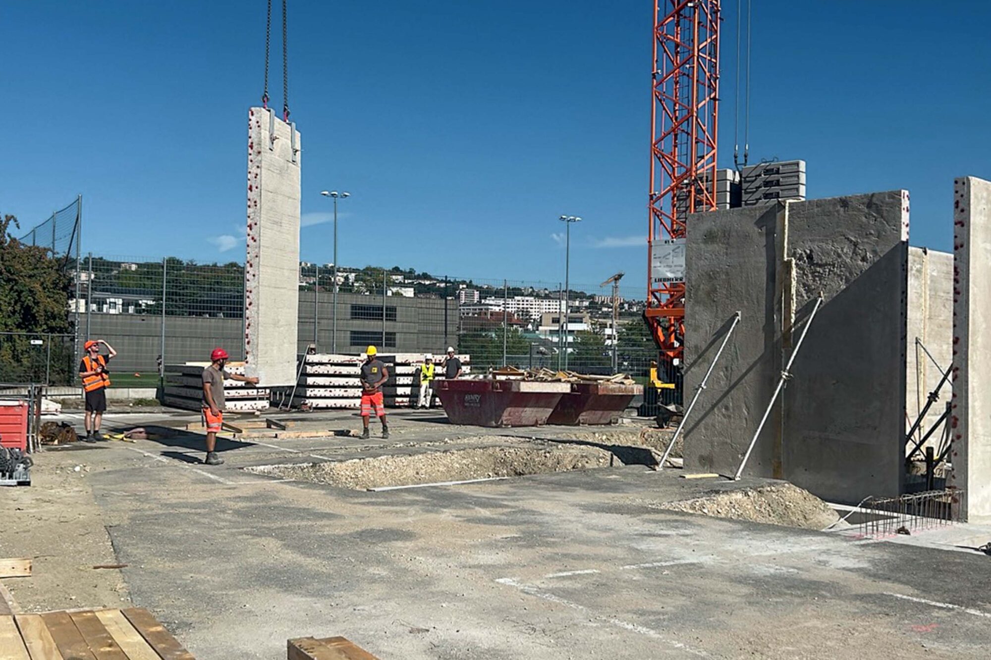 Une dalle de béton dressée à la verticale par la grue, au-dessus de murs déjà posés et d’ouvriers.