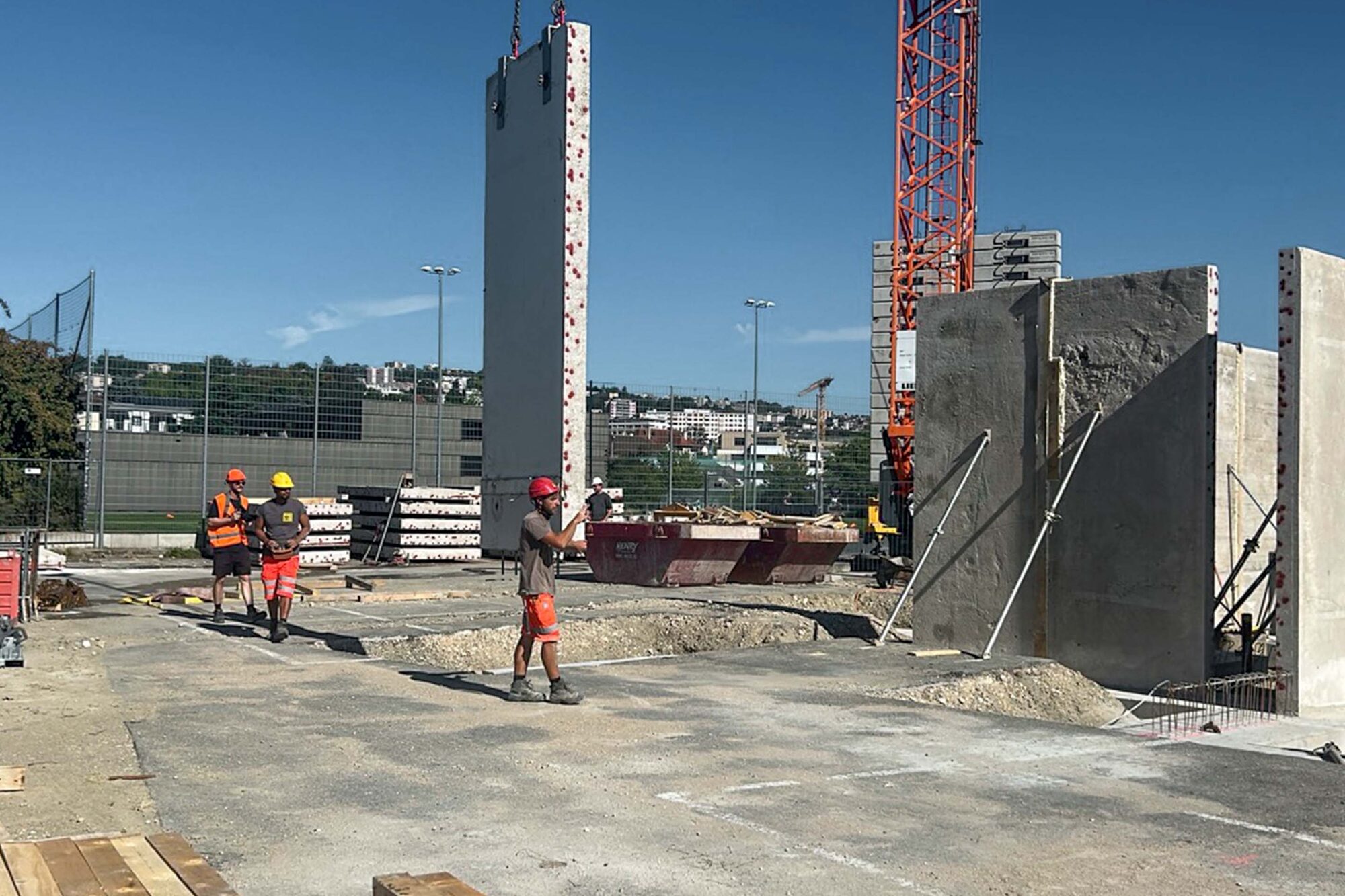 Une dalle de béton suspendue à la verticale, guidée par un ouvrier, à côté de murs déjà posés.