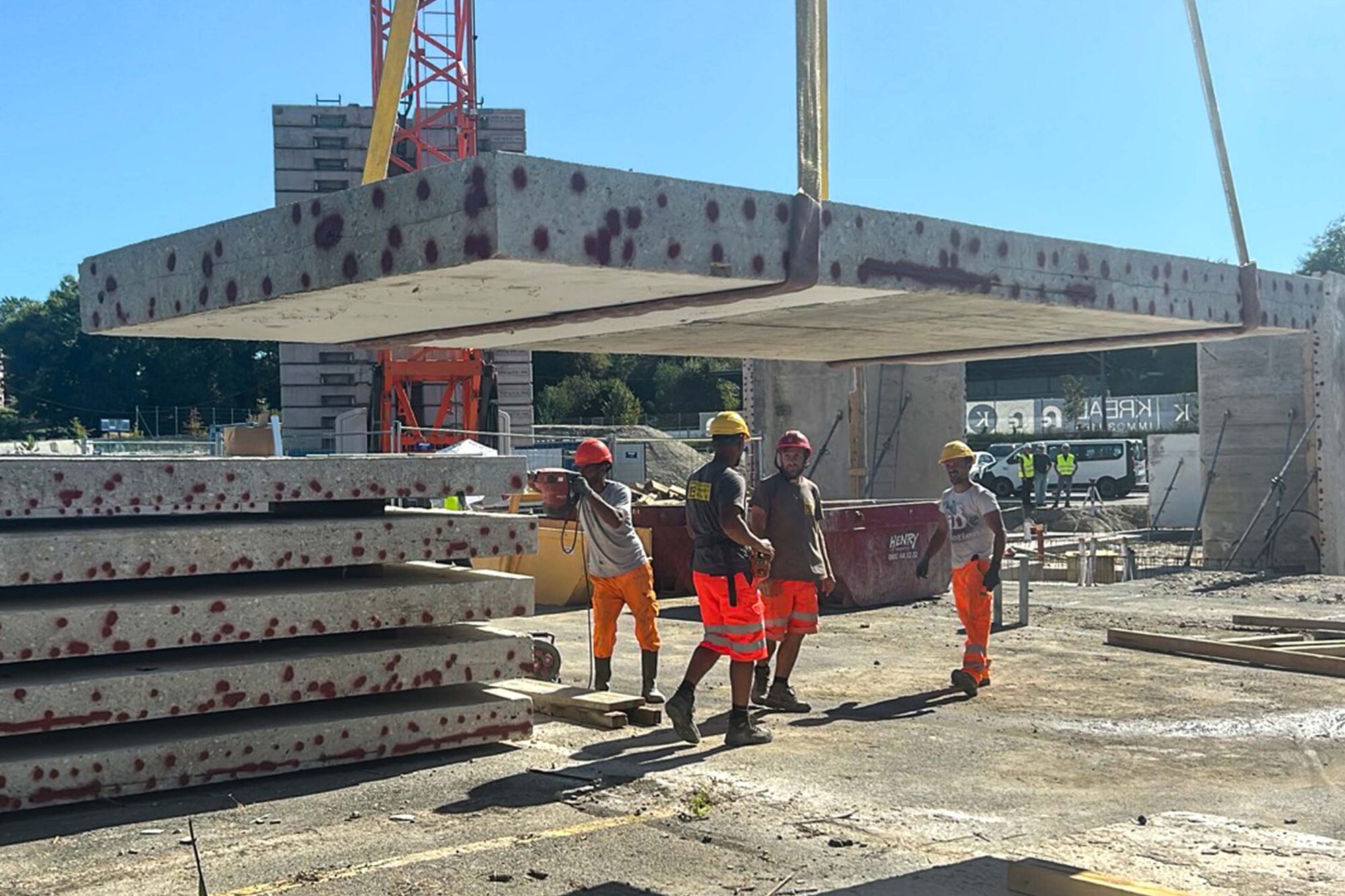 Dalles de béton empilées sur le chantier, avec une dalle suspendue à la grue et quatre ouvriers au premier plan.