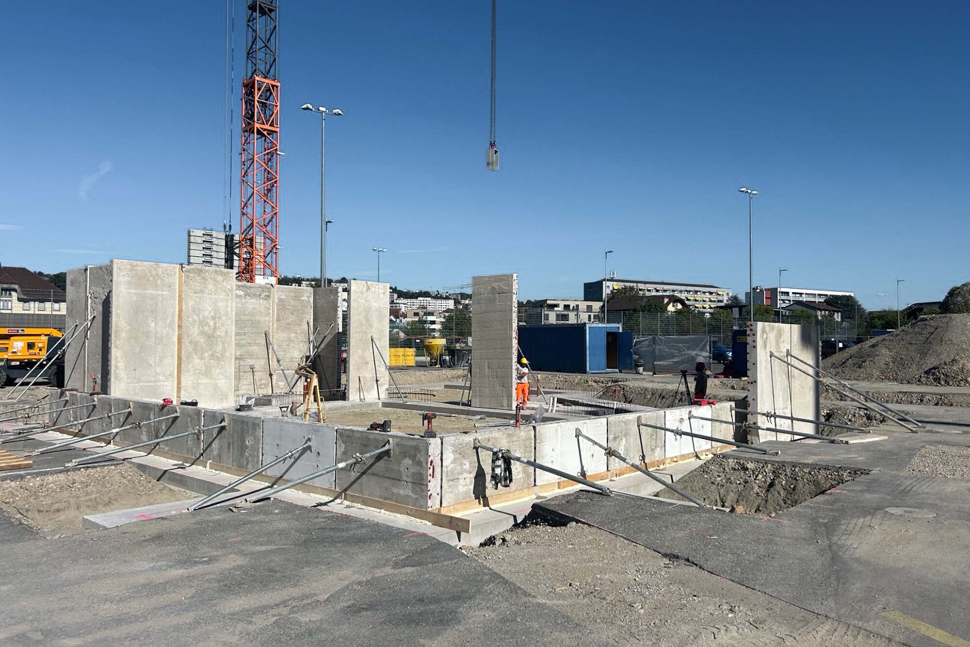 Murs de béton dressés sur une fondation linéaire, avec une grue et des maisons en arrière-plan.