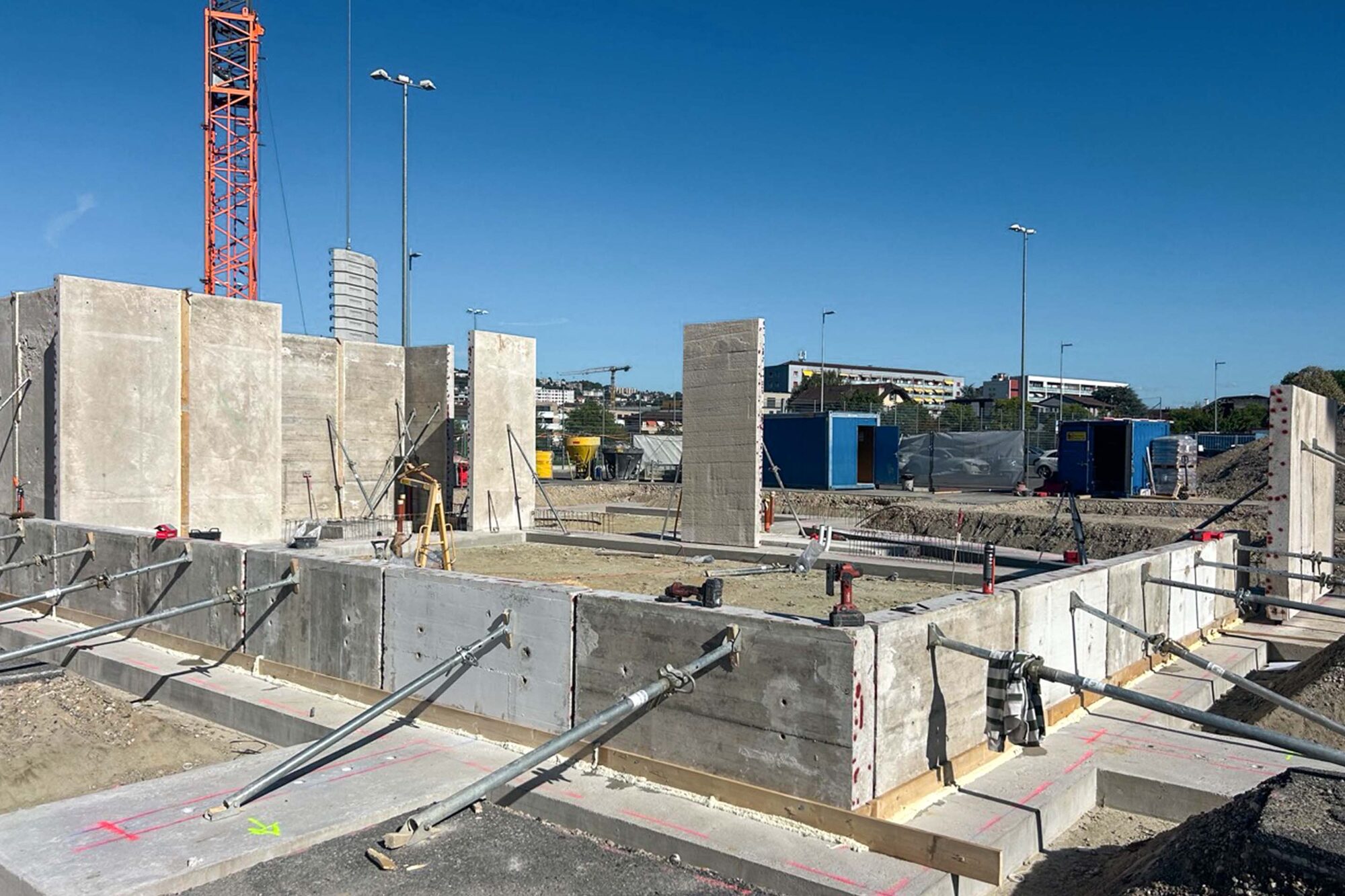 Vue d’ensemble du chantier avec murs de béton étayés et grue, depuis le bord de la fondation.