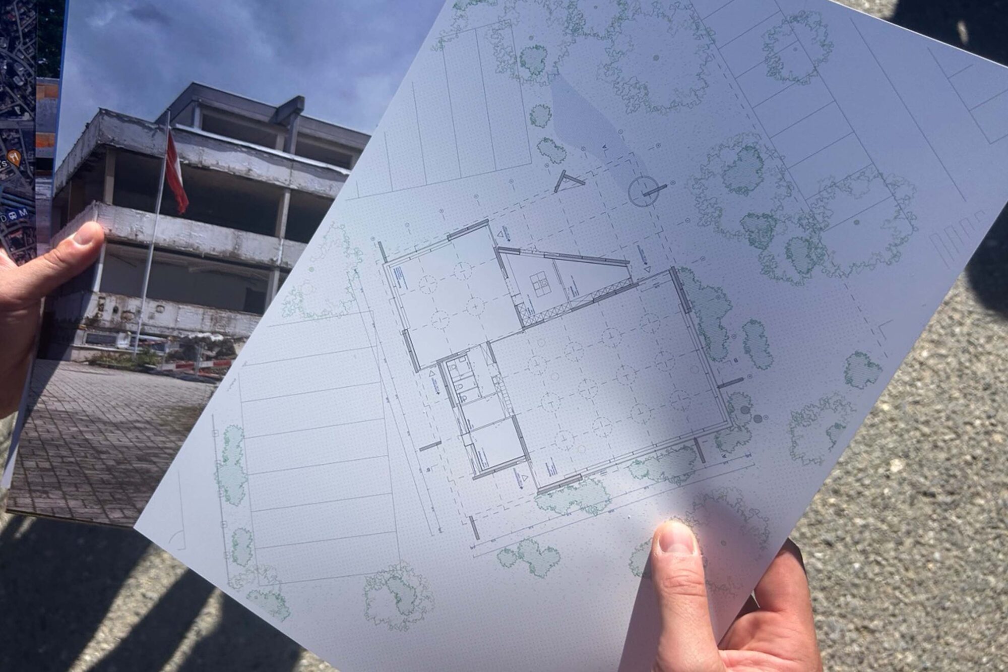Main tenant un plan d’implantation du bâtiment, devant un immeuble de bureaux.