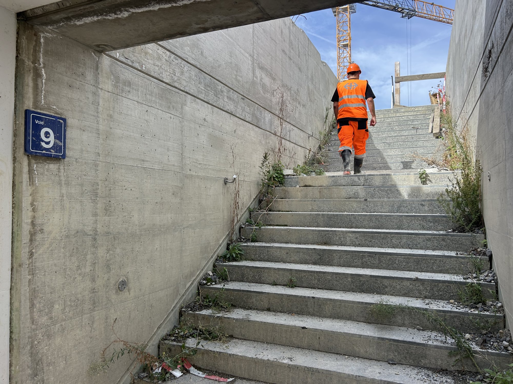 Homme en tenue orange montant un escalier en béton, plaque «Voie 9» à gauche.