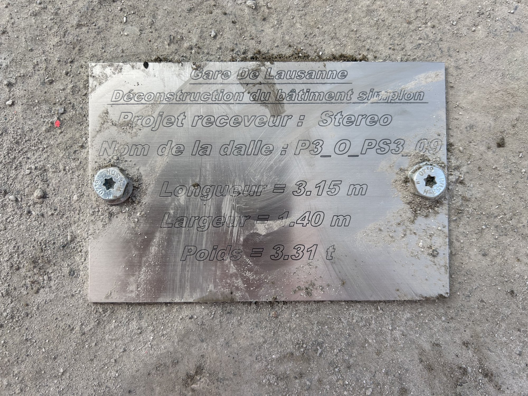 Plaque métallique gravée posée sur du béton, indiquant le projet receveur et le poids de la dalle.