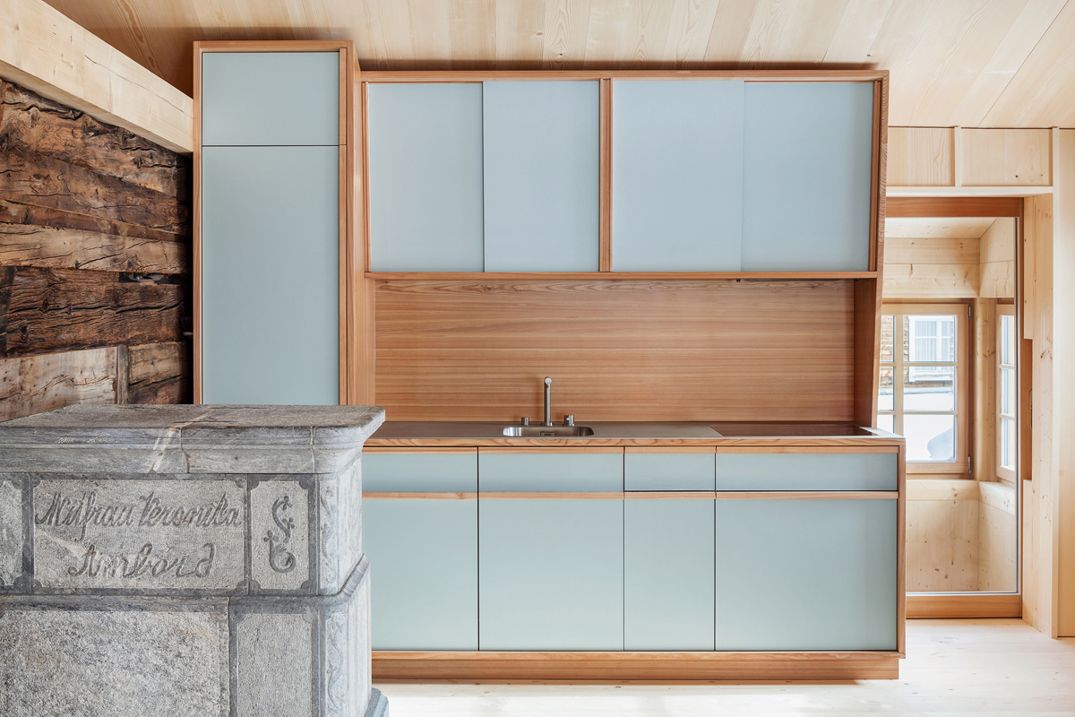 «Sonderpreis Energie»: Im Vordergrund ist einer der sanierten Giltsteinöfen zu sehen. Die neue Küche ist aus Lärche, der übrige Innenausbau ist in Fichtenholz ausgeführt, das einen Kontrast zu den freigelegten dunklen Blockbauwänden bildet. Deckleisten verleihen den neuen Holzflächen Vielschichtigkeit und Tiefe.