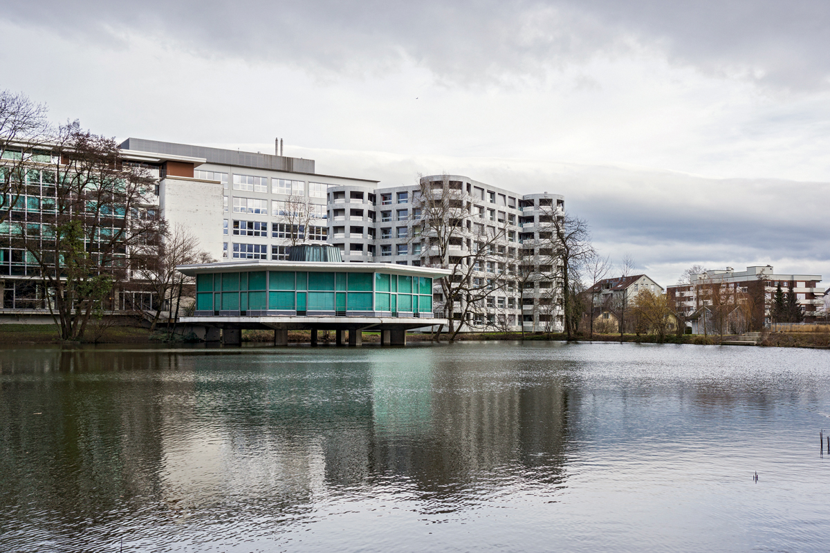Für die damalige Zellweger Uster AG baute der Architekt Roland Rohn das Verwaltungsgebäude mit dem Pavillon im Herterweiher. Heute blickt man über den Pavillon hinweg auf den Wohnbau von Herzog &amp; de Meuron.