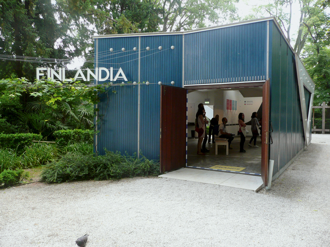 Ein Absperrband in den finnischen Nationalfarben begrüsst die Besucher im Türrahmen, um darauf hinzuweisen, dass man eine Grenze überschreitet und finnisches Hoheitsgebiet betritt. Dokumentiert werden die Ergebnisse eines Architekturwettbewerbs, bei dem Immigrantenunterkünfte konzipiert werden sollten.