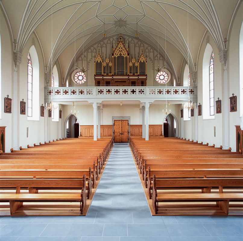 Die Raumhülle nach der Renovation 2014: Blick gegen die Empore. Die Dekorationsmalerei in beige-grauer Grisailletechnik vermittelt den Eindruck von aufwendigen Steinmetz- und Stuckaturarbeiten. Der Sakralraum wirkt hell und einladend.