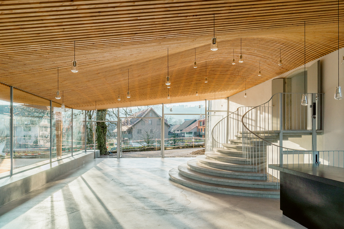 Im Innern wirkt das Foyer offen, und das 9 × 15 m messende Dach wölbt sich leicht über den neuen Treppenaufgang aus Mägenwiler Muschelkalk. Was wie ein leichtes Lattenwerk erscheint ist, in Wirklichkeit die Untersicht des Holztragwerks selber, gebildet aus 46 unterschiedlichen Trägerformen aus Brettschichtholz. Rechts die Holztheke mit Lackierung aus schwarz glänzendem Urushi, an der Decke angehängt die eigens entworfenen, mundgeblasenen Pendelleuchten.
