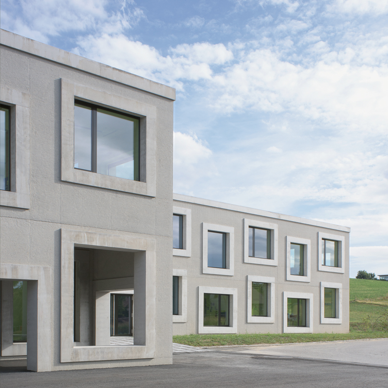 Die Fassaden der neuen Primarschule von Avry sind allseitig aus Beton mit gebrochenem Jurakies gefertigt. Das Aufrauen der Oberflächen mittels Sandstrahler liess den Kies hervortreten, was dem Bau einen warmen, erdigen Naturton verleiht – ein lebendiger Kontrast zum expressiv-farbigen Innenleben der Schule.