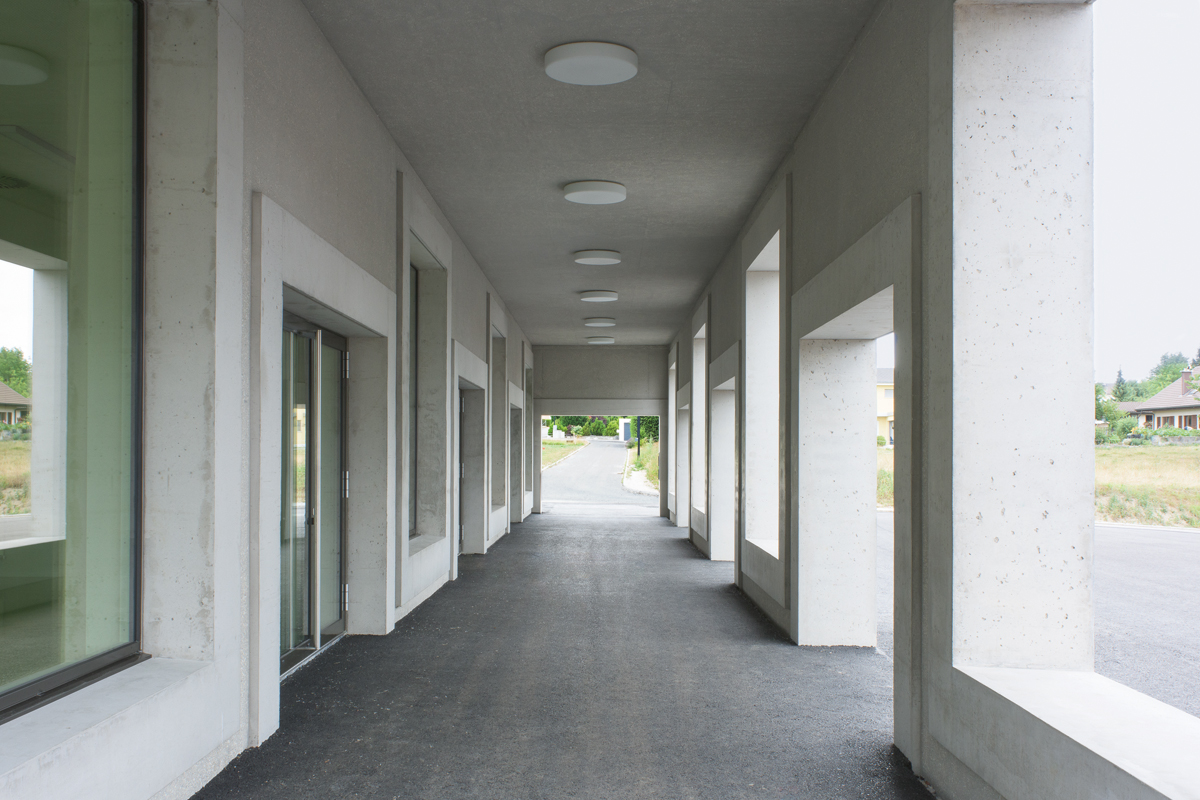 Vor den beiden Eingängen zu Saal und Schule ist die Fassade als Arkade ausgebildet. Der geschützte Aussenraum liegt in der ­Ver­län­ge­rung der Strasse, die ins angrenzende Wohngebiet führt. Die analog zu den Fenstern gestalteten Öffnungen können, wie die regulären Fensterbänke, als Sitzgelegenheiten genutzt werden.