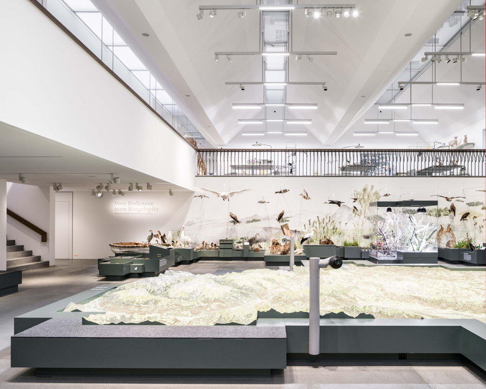 Der Raum mit dem Kantonsmodell bildet den Knotenpunkt der Ausstellung. Die Wandmalerei bindet ihn ein, von der Galerie lässt sich das ganze Modell überblicken, und über der Szenerie scheint das prägnante Dach zu schweben. Die das Tageslicht ergänzenden Leuchtkörper sind in die Architektur integriert.