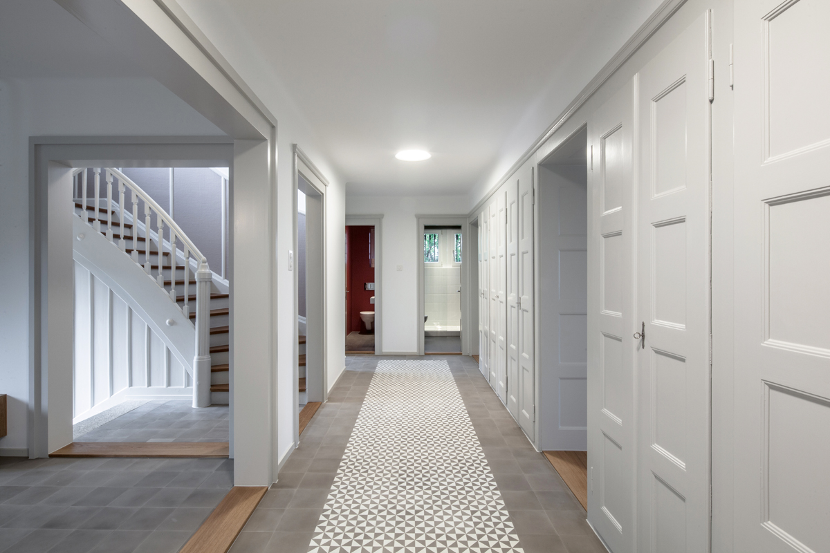 In der ehemaligen Küche befindet sich heute der neue Eingangsbereich. Wände zum Treppenhaus und zum Wohnungsflur wurden entfernt, die Bodenschwellen aber am Boden belassen, damit sich die Eingriffe deutlich abzeichnen. Das Entree ist mit grauen Zementplatten (mit einer darunter liegenden Bodenheizung) gestaltet und setzt sich im anschliessenden Korridor mit einer grafisch gemusterten Variante fort.