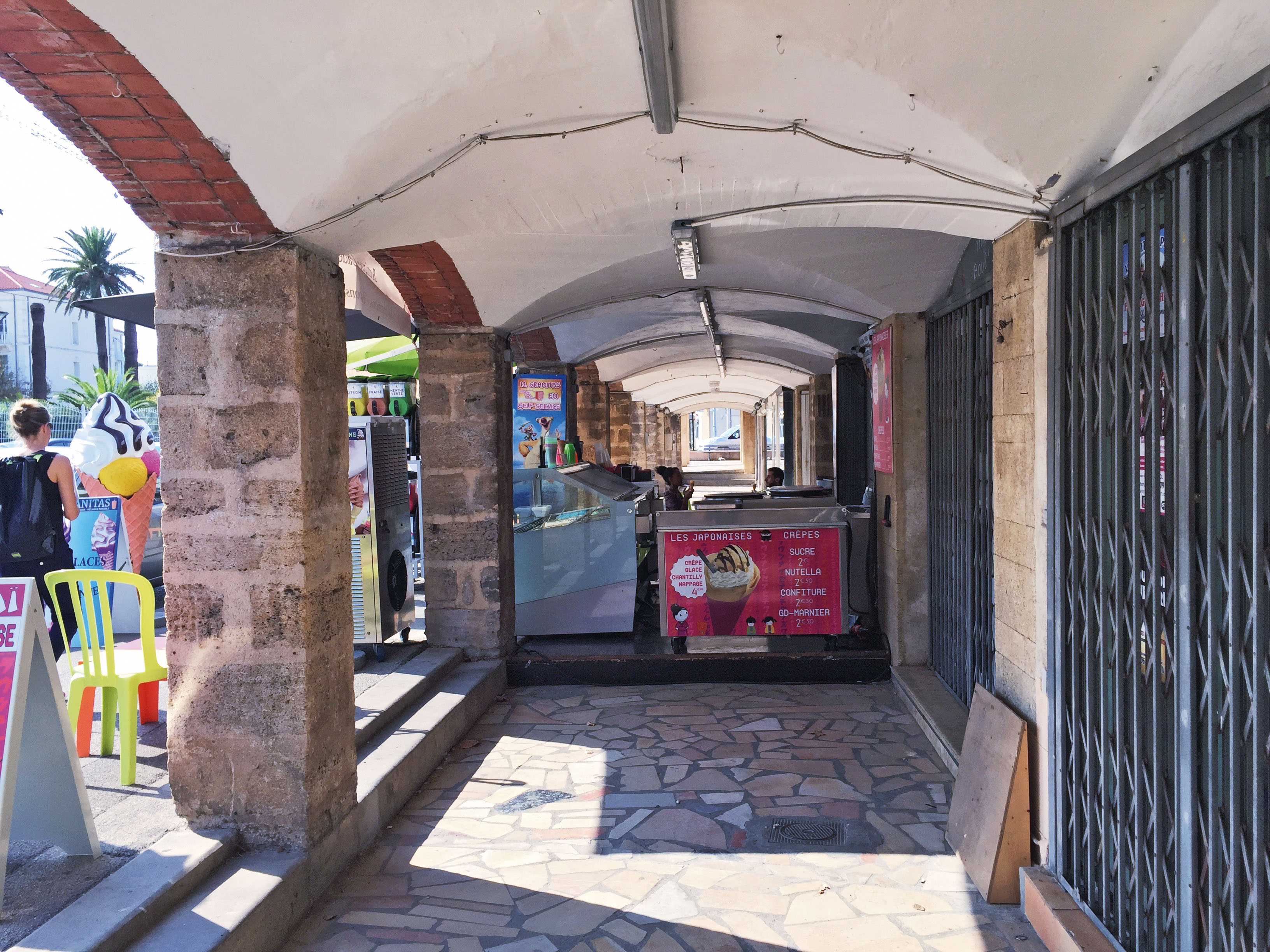 Dans la galerie marchande des boutiques nord, le passage sous arcades est obstrué par un commerce qui y déploie sa terrasse et ses comptoirs à glaces. On devine sous l’enduit les voûtes croisées en brique d’origine.