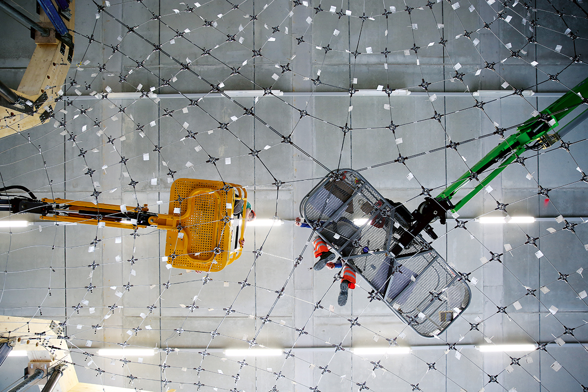 Arbeiter schweben auf Hebebühnen über dem Stahlkabelnetz, das – mit einer Membran überzogen, die hier nicht zu sehen ist – als Schalung für den Prototyp der Betonschale des HiLo dient.