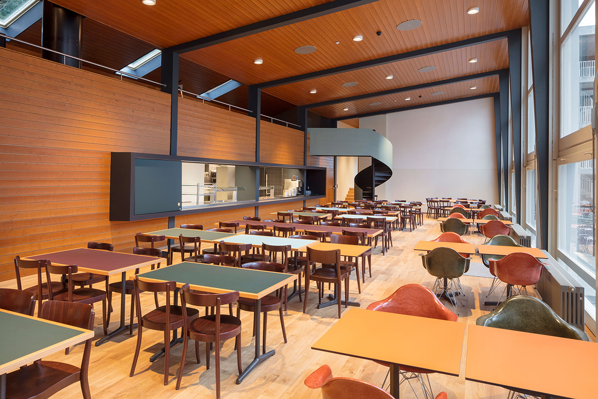 Der Saal öffnet sich mit einer grossen Glasfassade zum Platz. Hinter der Holzwand befindet sich die Küche. Das Mobiliar im Speisesaal wurde in Anlehnung an die Material- und Farbwelt des Bestands ausgewählt.