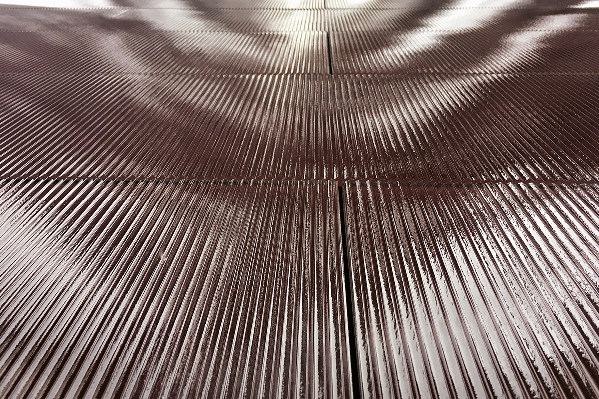 Die PV-Fassade am Haus «Solaris» in Zürich Wollishofen mit dem typischen vertikal verlaufenden Strukturen des Gussglases, dessen rötlicher Farbdruck die PV-Zellen versteckt. Das asymmetrische Profil des Glases ergibt Reflexionen, die an Lichtspiele auf einer Wasseroberfläche erinnern.