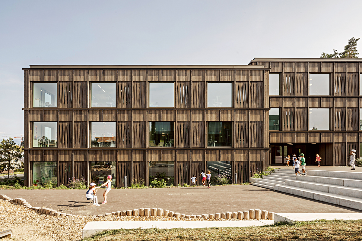 Die neue Primarschule von Nordwesten aus. Die Holzfassade verweist zum einen auf den benachbarten Wald, zum anderen aber auch auf die umliegenden Landwirtschaftsgebäude, die die Gemeinde noch heute prägen.