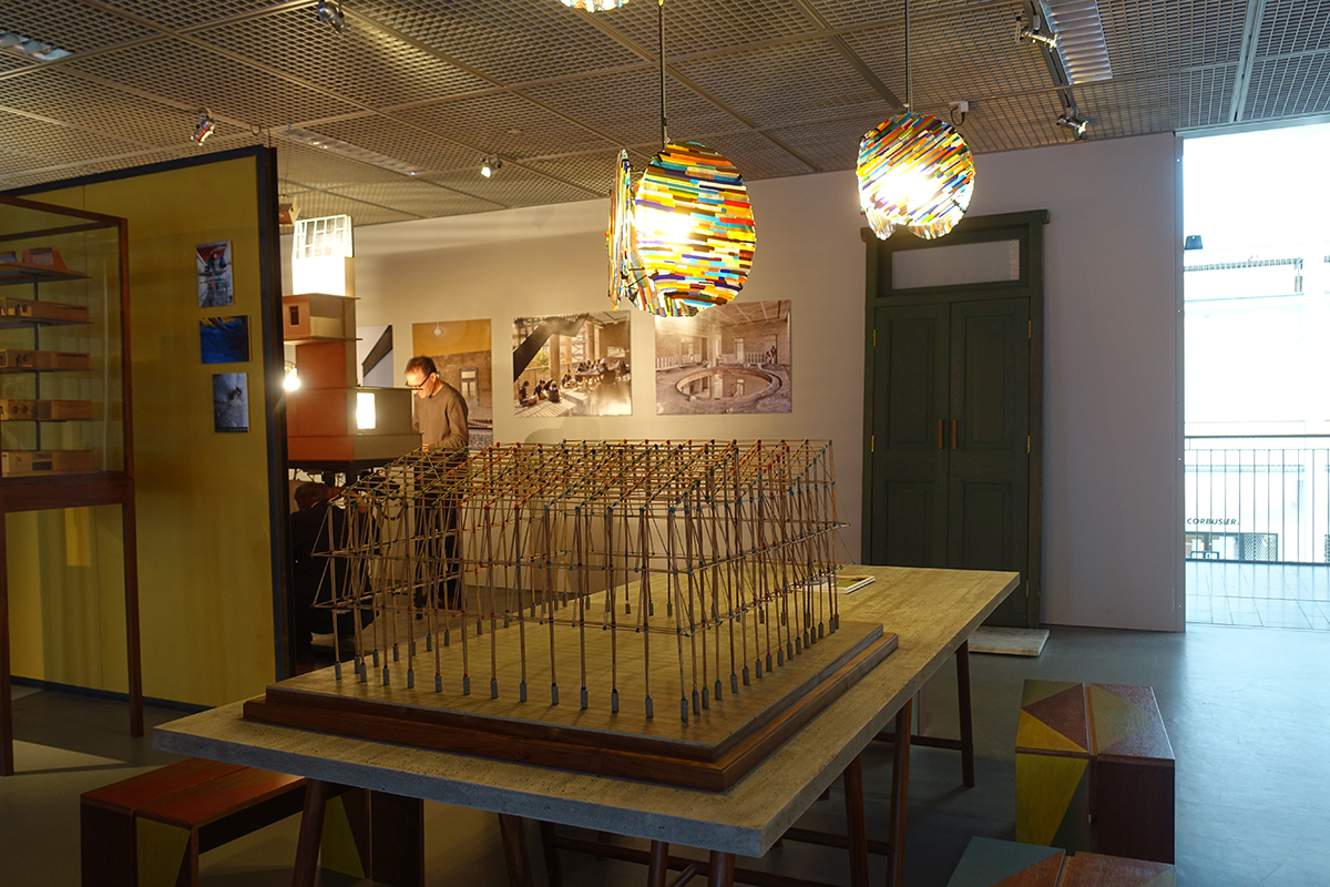 Blick in die Ausstellung «Case Design» bei Archizoom. Im Hintergrund Sam Barclay beim Einrichten des Modells zur solarbetriebenen Lüftung. Im Vordergrund ein Modell für die geplante Bambuskonstruktion einer Sporthalle. Die hängenden Leuchten sind während der Biennale in Zusammenarbeit mit venezianischen Glasbläsern entstanden. Reste von Buntglasstäben sind zu Platten verschmolzen, die Leuchten kommen später in die Bibliothek der Schule..