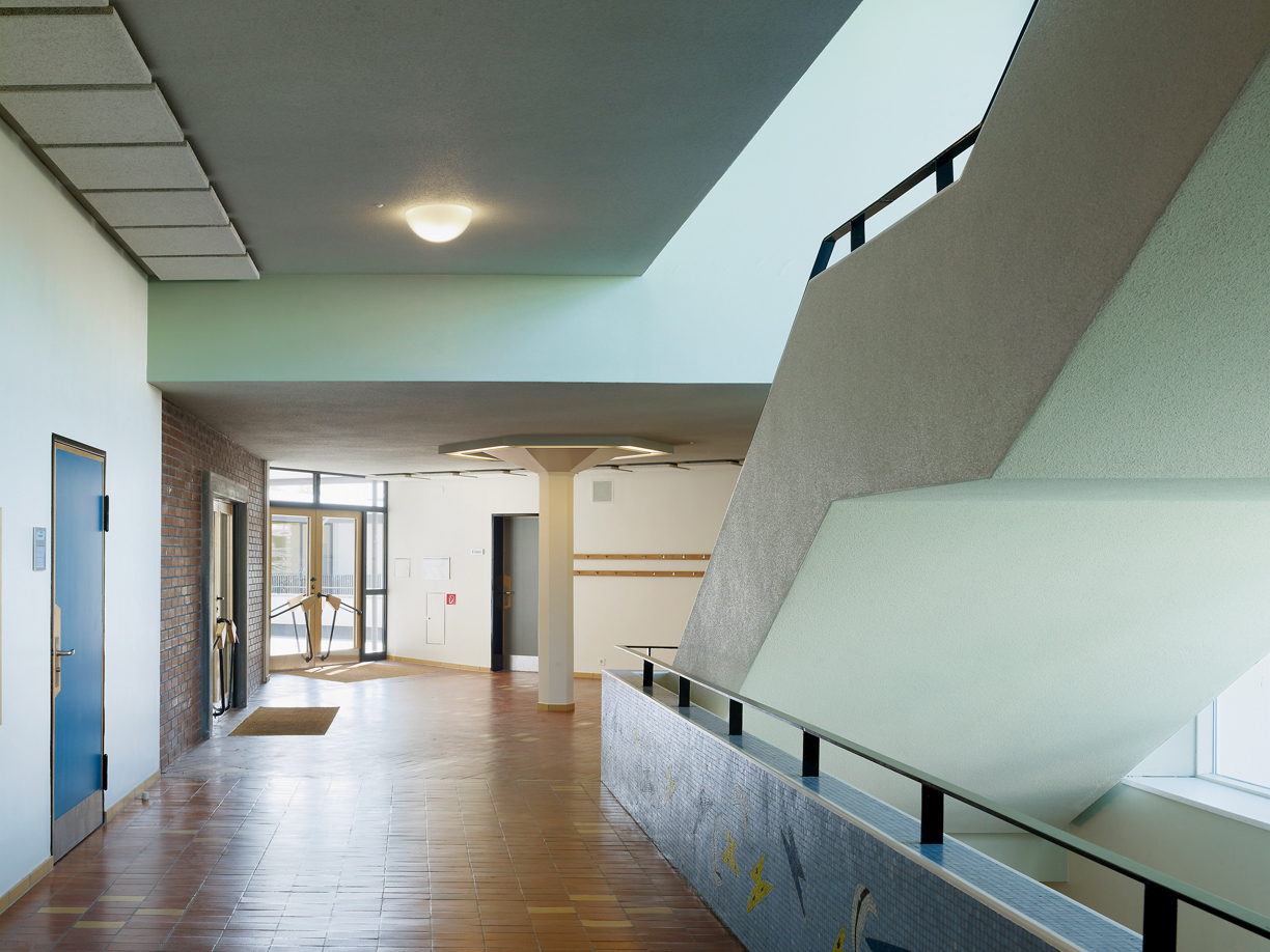 Seiteneingang der Schule (Trakt B) mit charakteristischer Pilzstütze und dem Treppenaufgang zum OG. Die Mosaiken von Charles Hindelang wurden aufgefrischt, ebenso die Böden aus gelbem und rotem Klinker. Neu erhöhen Brandschutzmassnahmen und eine Evakuationsanlage die Sicherheit. Die notwendige Unterteilung der Korridore wurde in der Haupterschliessungsachse mittels magnetauslösender Drehflügeltüren realisiert, die diskret in Wandnischen versteckt sind.