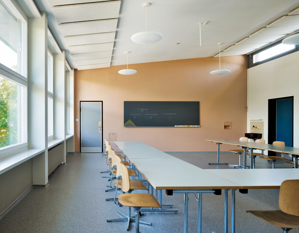 Klassenzimmer nach der Sanierung. Die bestehenden Akustikplatten wurden an der Fassadenseite um eine zusätzliche Reihe ergänzt, die Platten sind ein Nachbau im gleichen Stil. Bei den Beleuchtungskörpern setzten die Architekten auf eine Hybridlösung: Der Unterbau ist eine industriell gefertigte Pendelleuchte, die Befestigungsstäbe sind individuell beschichtet.