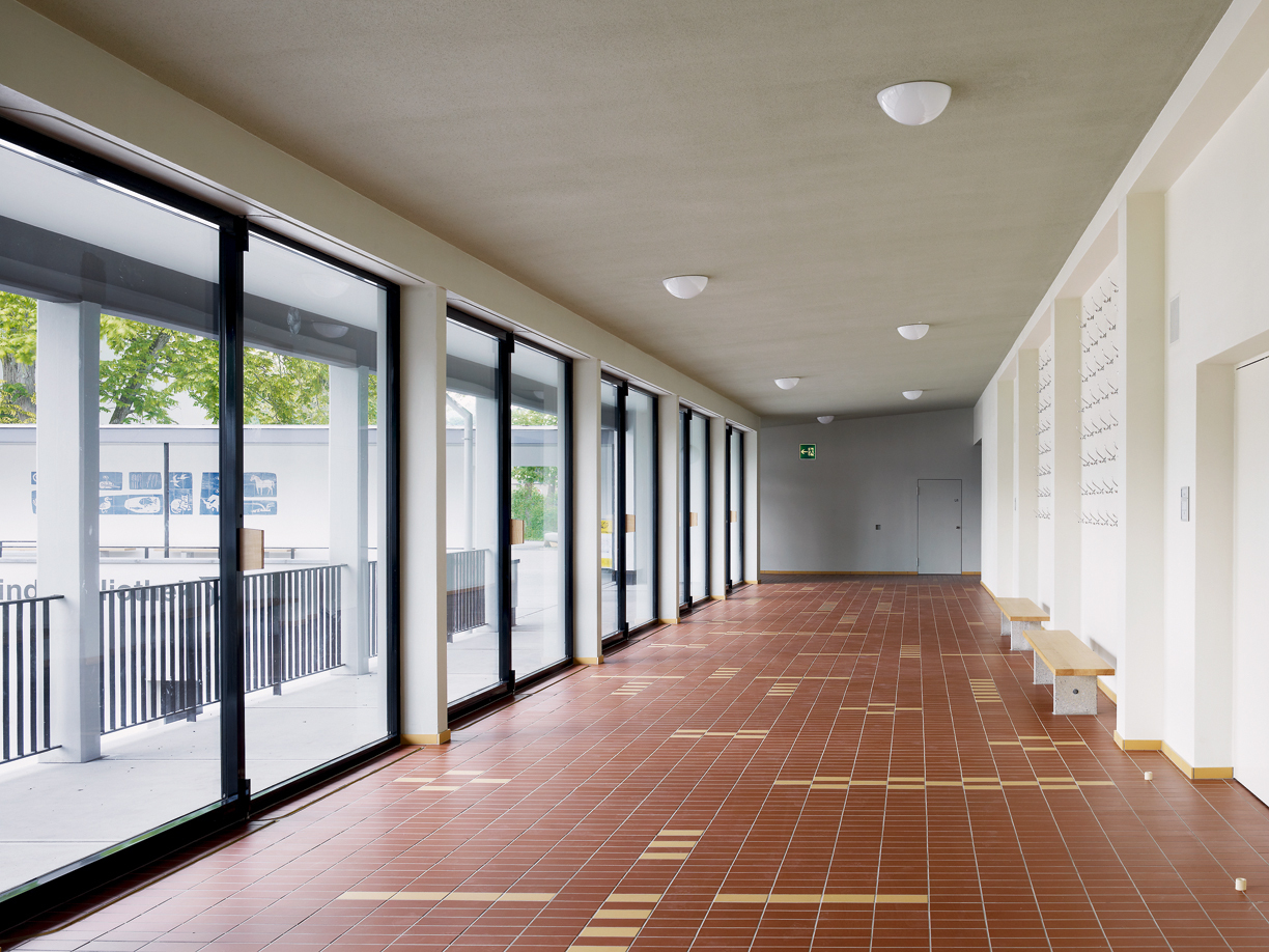 Das der Aula vorgelagerte Foyer: Die ehemals geschlossene Fassade wurden mit Glasschiebefenstern versehen. Wo früher die Umkleidekabinen untergebracht waren, öffnet sich der Bau nun zur Terrasse des Hauptzugangs. Es entsteht eine Blickachse zwischen Aula und Schulhaus, die räumliche Verbindung zwischen den Trakten wird gestärkt.